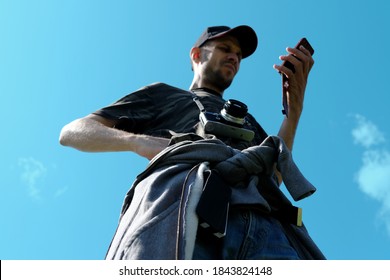 A man in a cap and with a camera around his neck looks at the phone against the sky. Bottom view.