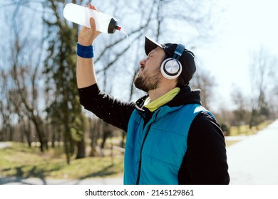 Man In A Cap And Headphones, An Athlete, A Runner In A Sleeveless Jacket Holds A Water Bottle He Is Dousing, Hot From The Sun, Spring Weather, A Sweaty Guy After A Marathon Run In The Park.