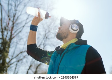 Man In A Cap And Headphones, An Athlete, A Runner In A Sleeveless Jacket Holds A Water Bottle He Is Dousing, Hot From The Sun, Spring Weather, A Sweaty Guy After A Marathon Run In The Park.