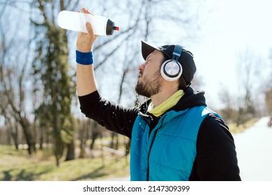 Man In A Cap And Headphones, An Athlete, A Runner In A Sleeveless Jacket Holds A Water Bottle He Is Dousing, Hot From The Sun, Spring Weather, A Sweaty Guy After A Marathon Run In The Park.