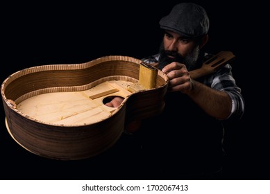 Man in cap, plaid shirt and apron checking guitar. Luthier manufacturing. Dark black background.