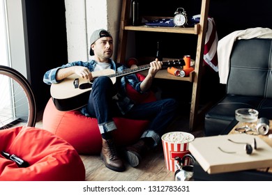 Man In Cap Playing Acoustic Guitar At Messy Living Room  