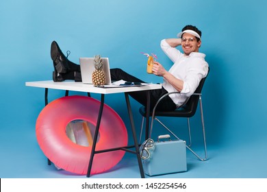 Man In Cap Works On Vacation And Drinks Cocktail. Guy Is Sitting At Table With Suitcase, Inflatable Circle, Laptop And Pineapple