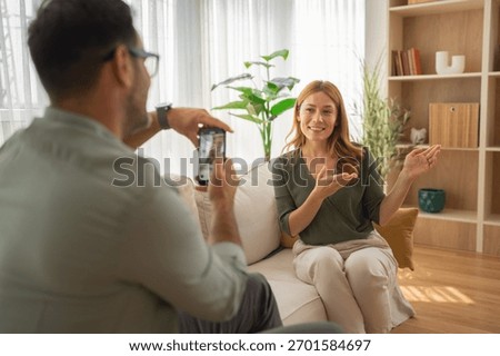 Man capturing video of a smiling influence woman actively presenting and communicating directly to a smart phone camera in a home living room, creating engaging content for online platforms