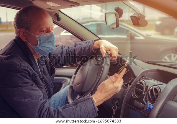 A man in a car with a\
phone in his hand and a protective mask against the spread of\
Coronavirus Covid-19