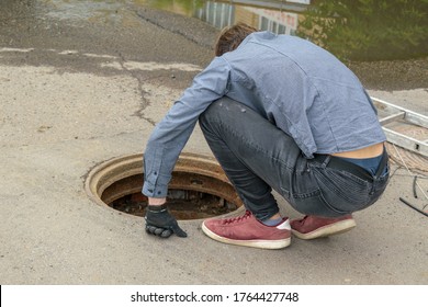 Man carefully looks at  open Manhole. Squatting bent over. View from the back. Concept of eliminating accident, laying communications, communication wires.