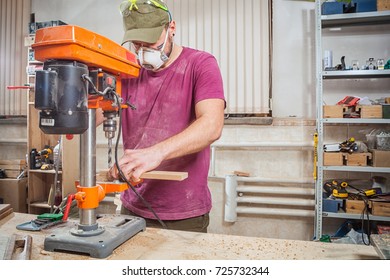 A Man Carpenter In A Cap, Safety Mask, Goggles And A T-shirt  Is Carving A Wooden Board On An Orange Large Drilling Machine In A Light Workshop Side View, In The Background A Lot Of Tools