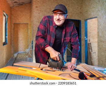 Man Is Carpenter. Guy With Electric Jigsaw Is Engaged In Woodworking. Carpenter Smiles And Looks Into Camera. Man Carves Wooden Elements. Carpenter At Work. Worker Inside House Under Construction