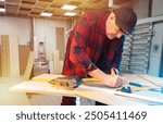 Man carpenter at work. Guy who makes furniture is standing in his workshop. Carpenter makes marks on boards. Man furniture maker in checkered shirt. Carpenter working in production workshop