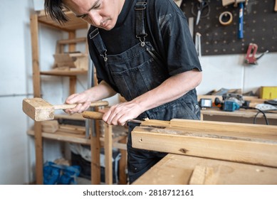 Man carpenter working at workbench with instruments and old wooden board in restoration workshop, concentrated master using chisel and mallet to cut off damaged part, preparing piece for patch.
