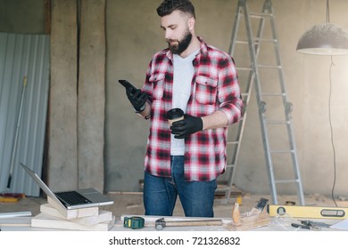 Man is carpenter,builder,designer stands in workshop, holds cup of coffee and uses smartphone. On desk is laptop and construction tools, in background a stepladder. Repair, construction, carpentry. - Powered by Shutterstock