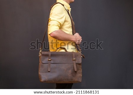 Man carries brown leather messenger bag in the hand on gray background. Unisex bag for sale.