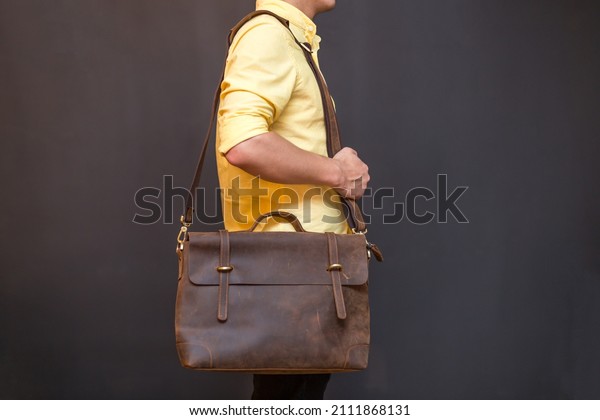 Man carries brown leather messenger\
bag in the hand on gray background. Unisex bag for\
sale.