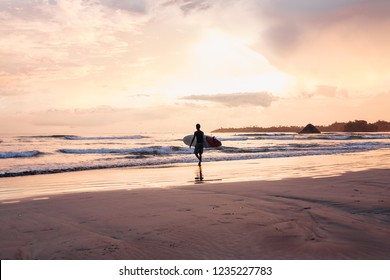 A Man Carries Surf On The Ocean On The Sunset. Guy Walking With Surfboard On The Beach.