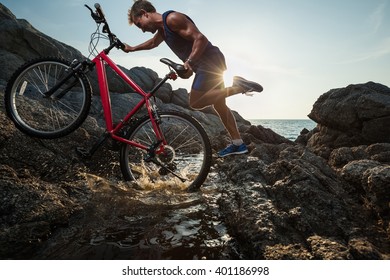 Man carrying a bike on the rock through water during sunset