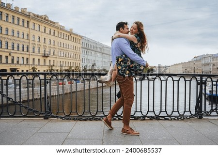 Man carrying a woman in his arms, both smiling, by the riverside railing. Guy with girlfriend at vacation trip, travel, honeymoon, romance.