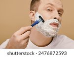 A man in casual attire carefully shaves his face with a razor.