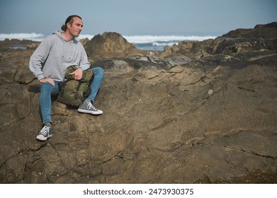 A man in casual attire sits on a rocky coastline with a green backpack, looking at the ocean waves. The scene captures a moment of relaxation and contemplation by the sea. - Powered by Shutterstock