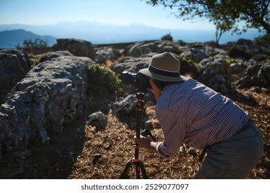 A man in casual attire uses a camera on a tripod to capture nature in a rocky terrain. The scene conveys exploration and a passion for photography amidst natural beauty. - Powered by Shutterstock