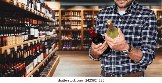 Man in casual clothes choosing wine in liquor store. Male customer holds two wine bottles in hands while standing in wine shop. - Powered by Shutterstock