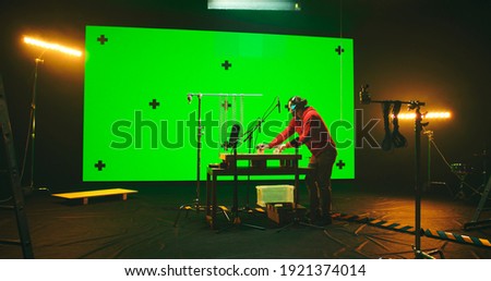 Man in casual clothes recording sounds on table under microphones against large screen with chromakey in dark studio