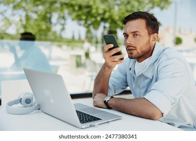 Man in casual clothing using a smartphone for voice command while working on a laptop outdoors. - Powered by Shutterstock