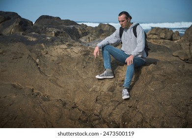 A man in a casual grey hoodie and jeans, with a backpack, sitting on a rocky shore by the beach. He appears contemplative, enjoying a peaceful moment by the ocean on a sunny day. - Powered by Shutterstock