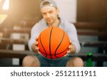 A man in casual sportswear sits on outdoor bleachers, holding a basketball directly toward the camera. The focus is on the basketball, with the man