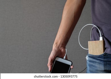 A Man In Casual Wear Holding Smartphone Charging With Power Bank On Grey Wall Background, Technology And Lifestyle Concept.