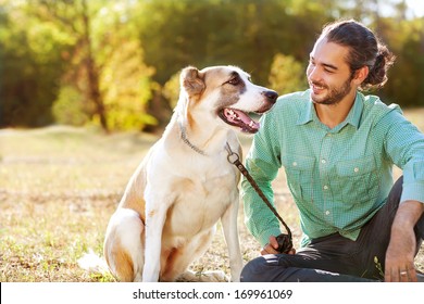 Man And Central Asian Shepherd Walk In The Park. He Keeps The Dog On The Leash.