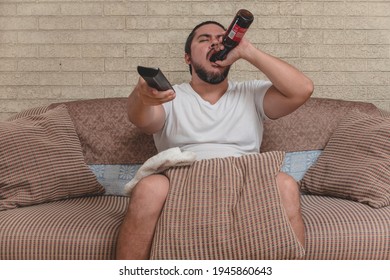 A man changes channels or chooses from menu option with a remote while chugging down a bottle of beer and sitting on the couch. Slacker and drunkard.