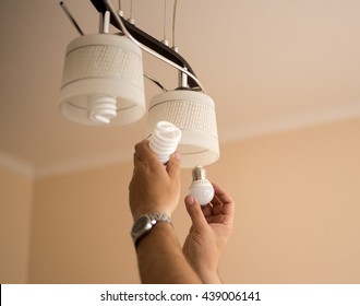 Man Changes An Electric Led Light Bulb, Energy Efficiency. Closeup Of Man's Hand Adjusting Electric Bulb By Pendant Lights At Home.