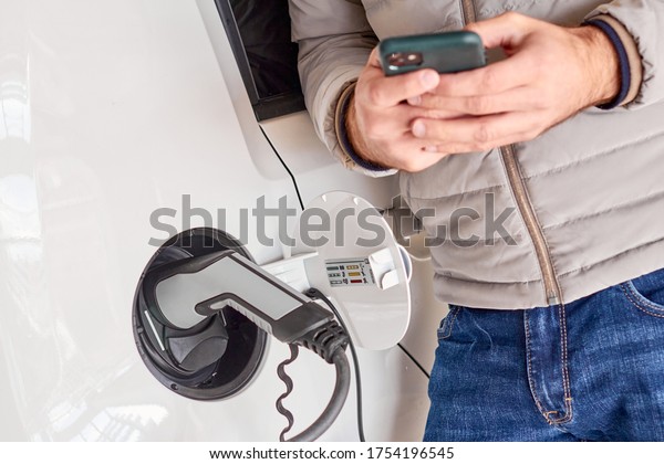 Man charging electric automobile and\
using his smartphone for payment at charging\
point.