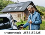 Man charging electric car in front of his house, plugging the charger into the charging port. House with solar panel system on roof behind him.