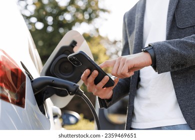 Man charging electric car at charging station using smart phone app. Unrecognizable male unplugging electric car from charging station. Man is unplugging in power cord to an electric car at sunset. - Powered by Shutterstock