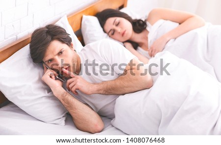 Man cheater talking privately on cellphone in family bed, showing hush sign while his wife sleeping, panorama