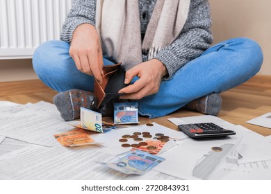 Man checking empty wallet sitting on floor near radiator with bills and money, struggling with heating costs and debt. High quality photo