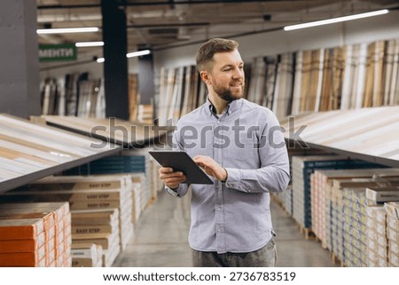 Man checking inventory system on a tablet in a retail warehouse, surrounded by stacked flooring products and samples