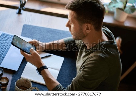 Man checking smartphone with unknown caller screen at desk with laptop and notebook. Incoming call disrupting person at workplace. Concept of mobile communication, scam call and distraction