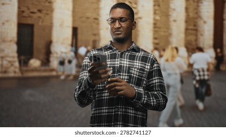 Man checking smartphone while holding a coffee cup on a historic street with stone columns and passersby; concern focus.