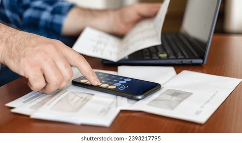 Man checking utility bills, taxes, bank account balance and calculates the amount using mobile phone sitting in the living room at home - Shutterstock ID 2393217795