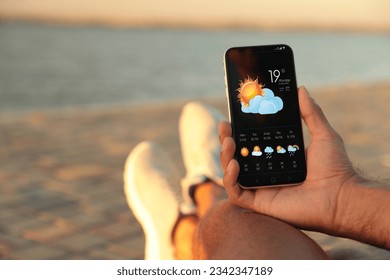 Man checking weather using app on smartphone near river, closeup. Data, sun with cloud and other illustrations on screen