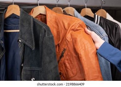 Man Checks Orange Leather Jacket In A Second Hand Store. Various Vintage Jackets Hang On Clothing Rack. Thrifting And Sustainability In Clothing Concept