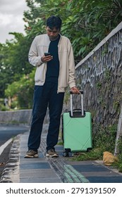 Man checks phone while pulling mint suitcase beside stone wall. He stands still, focused, on narrow outdoor path. Greenery climbs textured stone, adding natural charm. Ideal for travel, urban commute