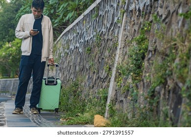 Man checks phone while pulling mint suitcase beside stone wall. He stands still, focused, on narrow outdoor path. Greenery climbs textured stone, adding natural charm. Ideal for travel, urban commute