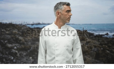 Man in chef jacket looking right on rocky coast with ocean and waves behind, subtle head turn and open mouth gesture; contemplation.