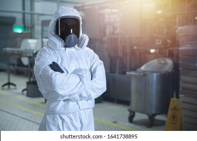 Man In Chemical Protection Clothes And Toned Mask In A Factory Against The Background Of An Industrial Workshop Looking Into The Frame. 