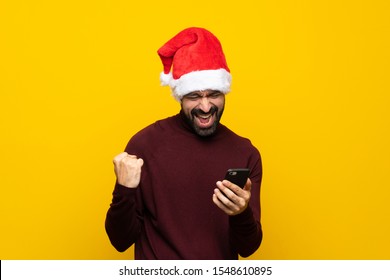 Man with christmas hat over isolated yellow background with phone in victory position - Powered by Shutterstock