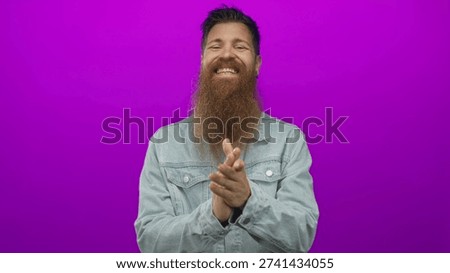 Man clapping hands in studio with a vibrant purple backdrop, wearing denim jacket and full beard; celebration appreciation gratitude joy.