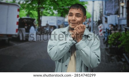 Man clasping hands and smiling in light jacket on urban street, goatee visible and hands held near chest facing camera; surprise curiosity.